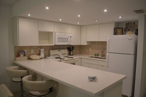 A modern kitchen with white cabinets, a built-in microwave, a refrigerator, and a bar area with two bar stools.