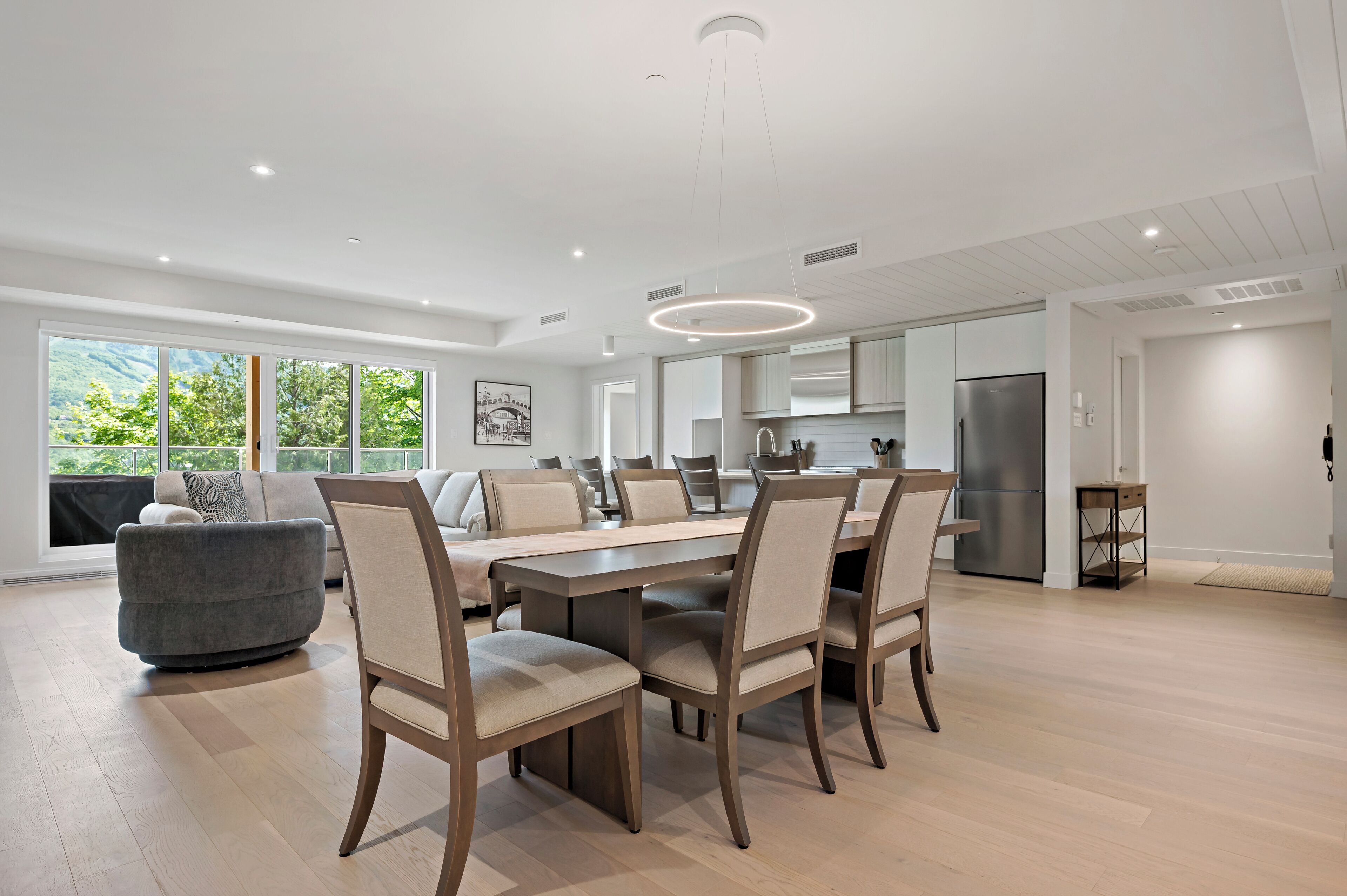 Un vaste espace dédié à la salle à manger, avec une longue table et des chaises, une cuisine moderne dotée d’un réfrigérateur en acier inoxydable et de grandes fenêtres offrant une vue sur la verdure.