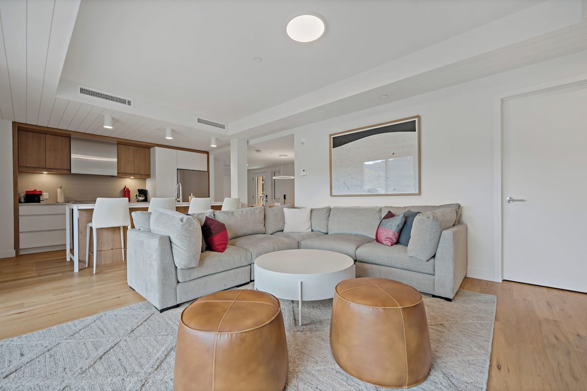 Een moderne woonkamer met een grijze hoekbank, een witte salontafel en twee leren poefs. In de achtergrond is een keuken met witte kasten en een eethoek met witte stoelen zichtbaar.
