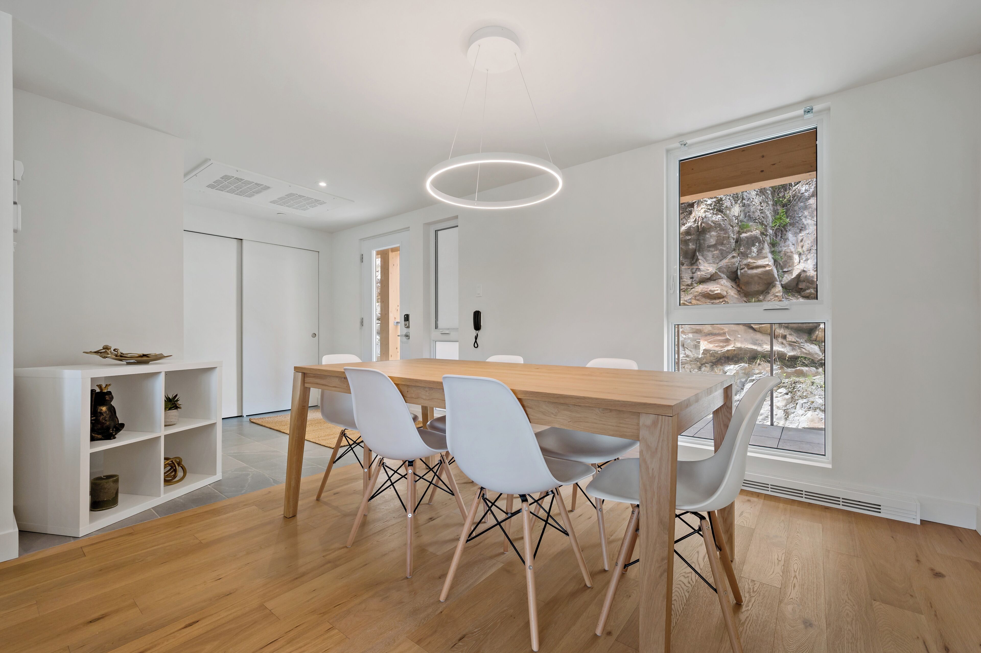 A modern dining area with a wooden table and white chairs, a white shelving unit, and a large window with a view of rocks.