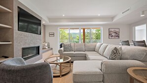 A modern living room with a large sectional sofa, a flat-screen TV mounted on the wall, a fireplace, and large windows offering a view of greenery.