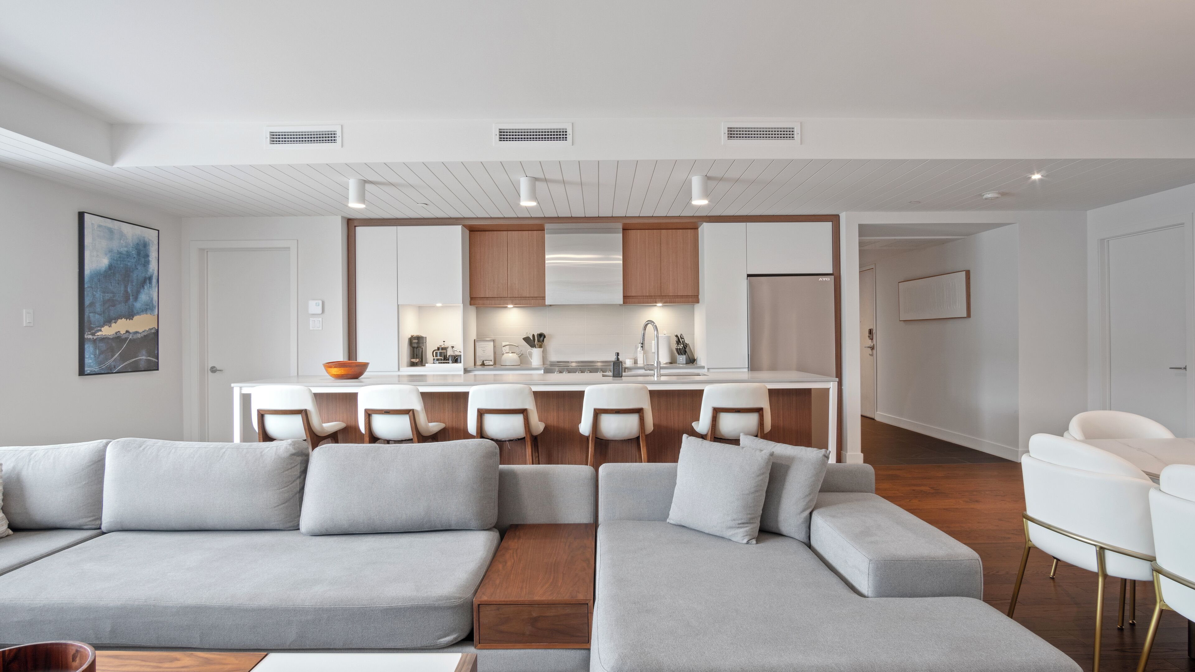 A modern living room with a grey sectional sofa, a wooden coffee table, and a kitchen area with a central island and bar stools.