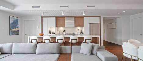 A modern living room with a grey sectional sofa, a wooden coffee table, and a kitchen area with a central island and bar stools.