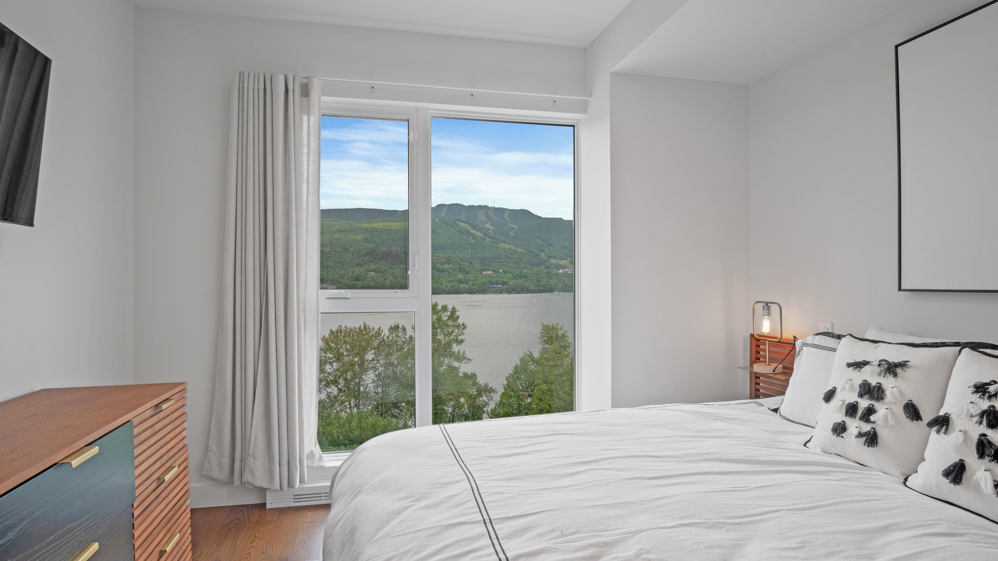 Une chambre avec une grande fenêtre, un lit recouvert de draps blancs, une table de chevet avec une lampe, et une vue sur un lac et des collines.