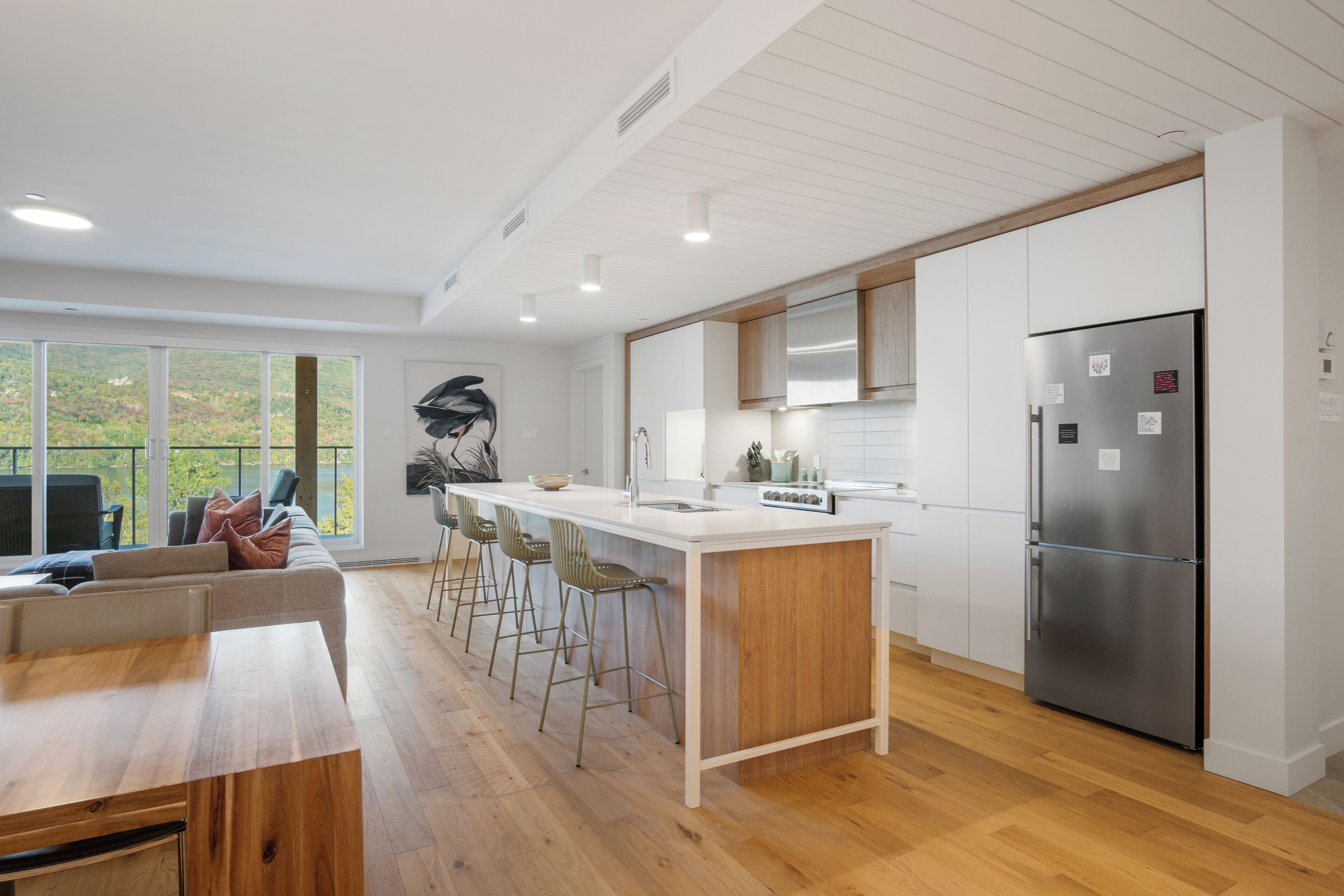 Una cocina moderna con una isla central, taburetes de bar, un refrigerador de acero inoxidable y vistas al exterior a través de grandes ventanales.