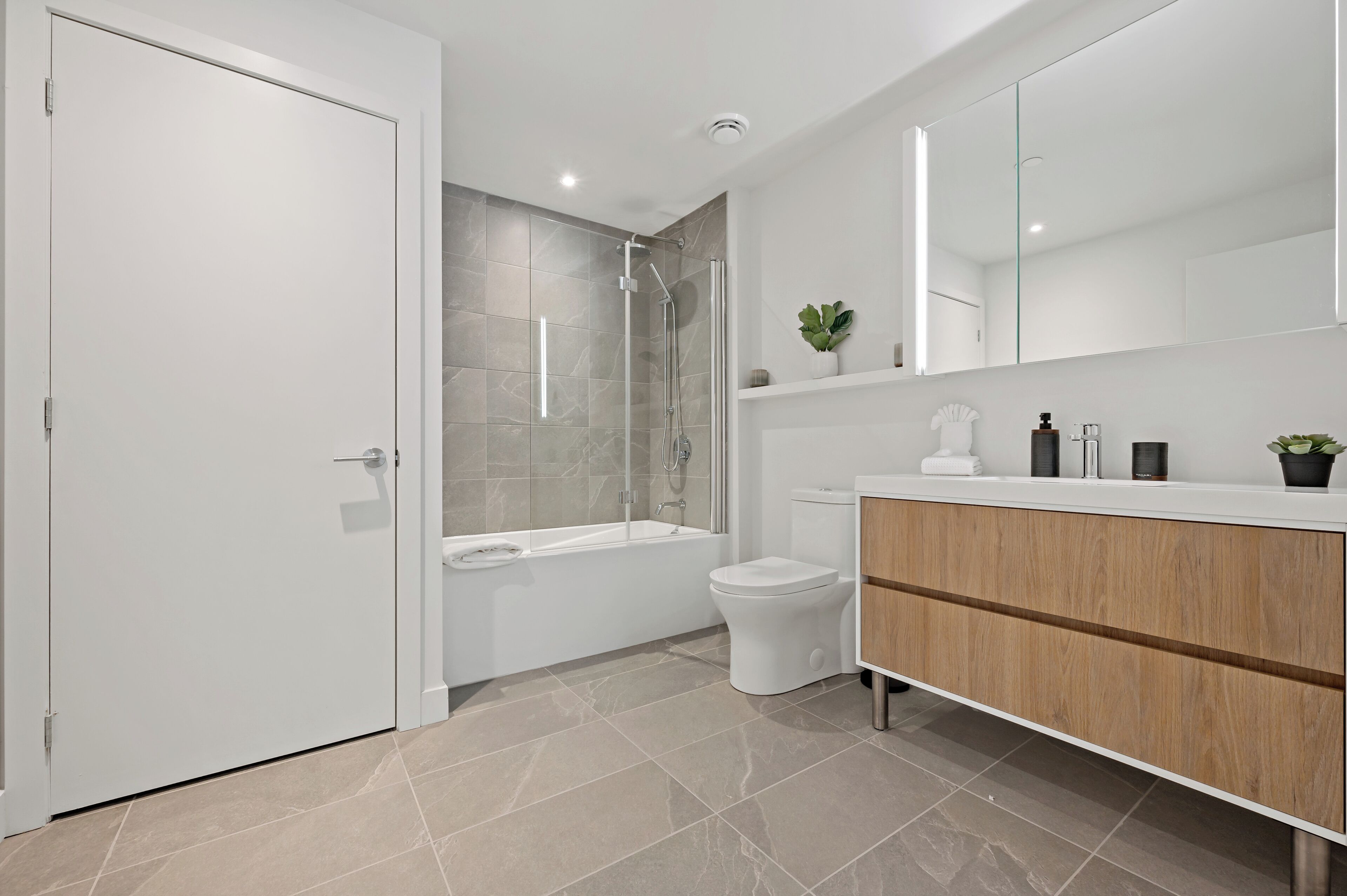 Une salle de bain moderne avec une porte blanche, une douche en verre, une baignoire blanche, des toilettes et un meuble-lavabo en bois avec un miroir.