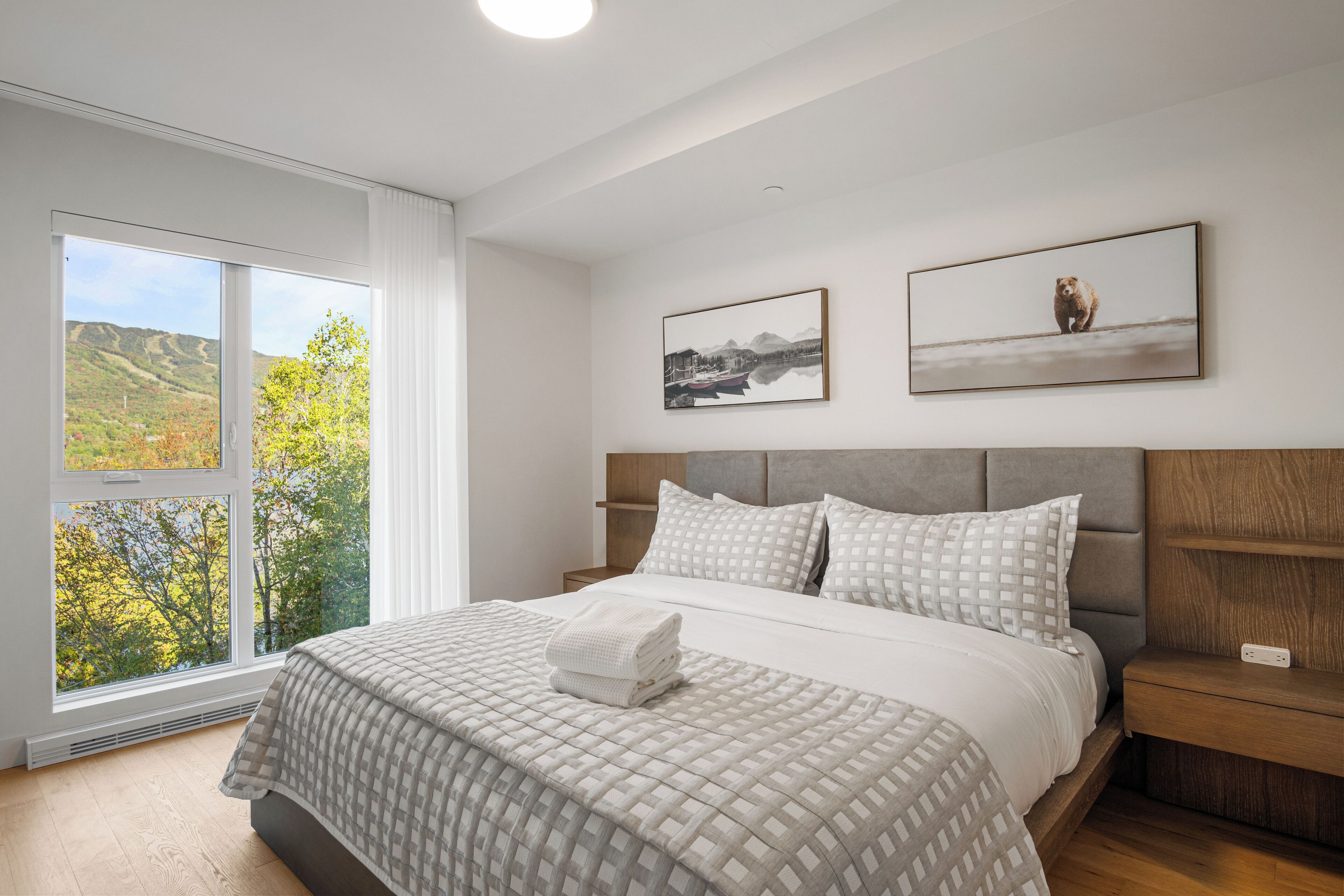A bedroom with a large window, a bed with a patterned bedspread, two wall-mounted pictures, and a wooden nightstand.