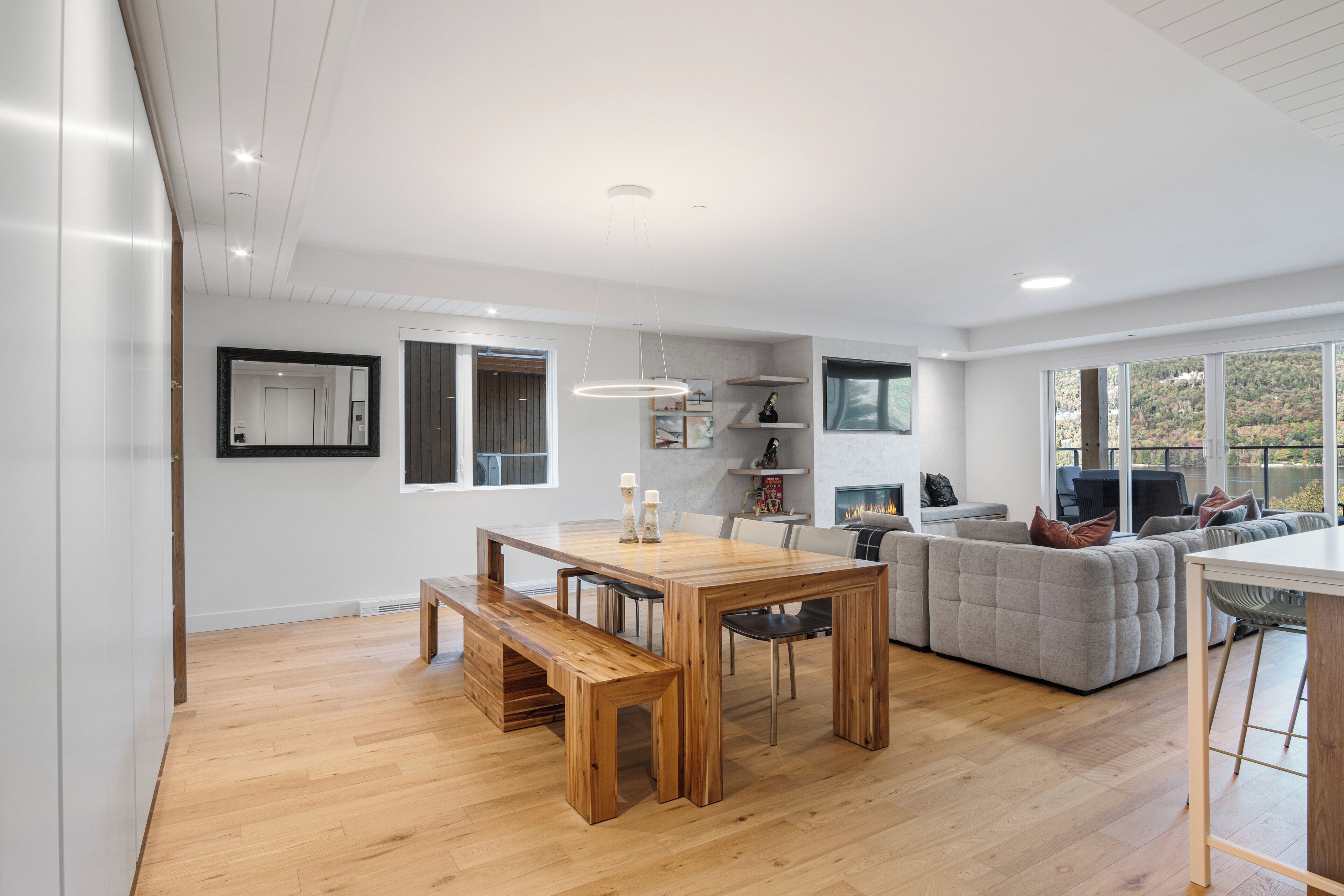 A modern living space with a wooden dining table, bench, grey sofa, and a white kitchen area.