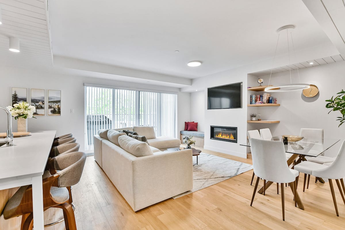 Een moderne woonkamer met een bank, eettafel en stoelen, een open haard en een aan de muur bevestigde televisie.