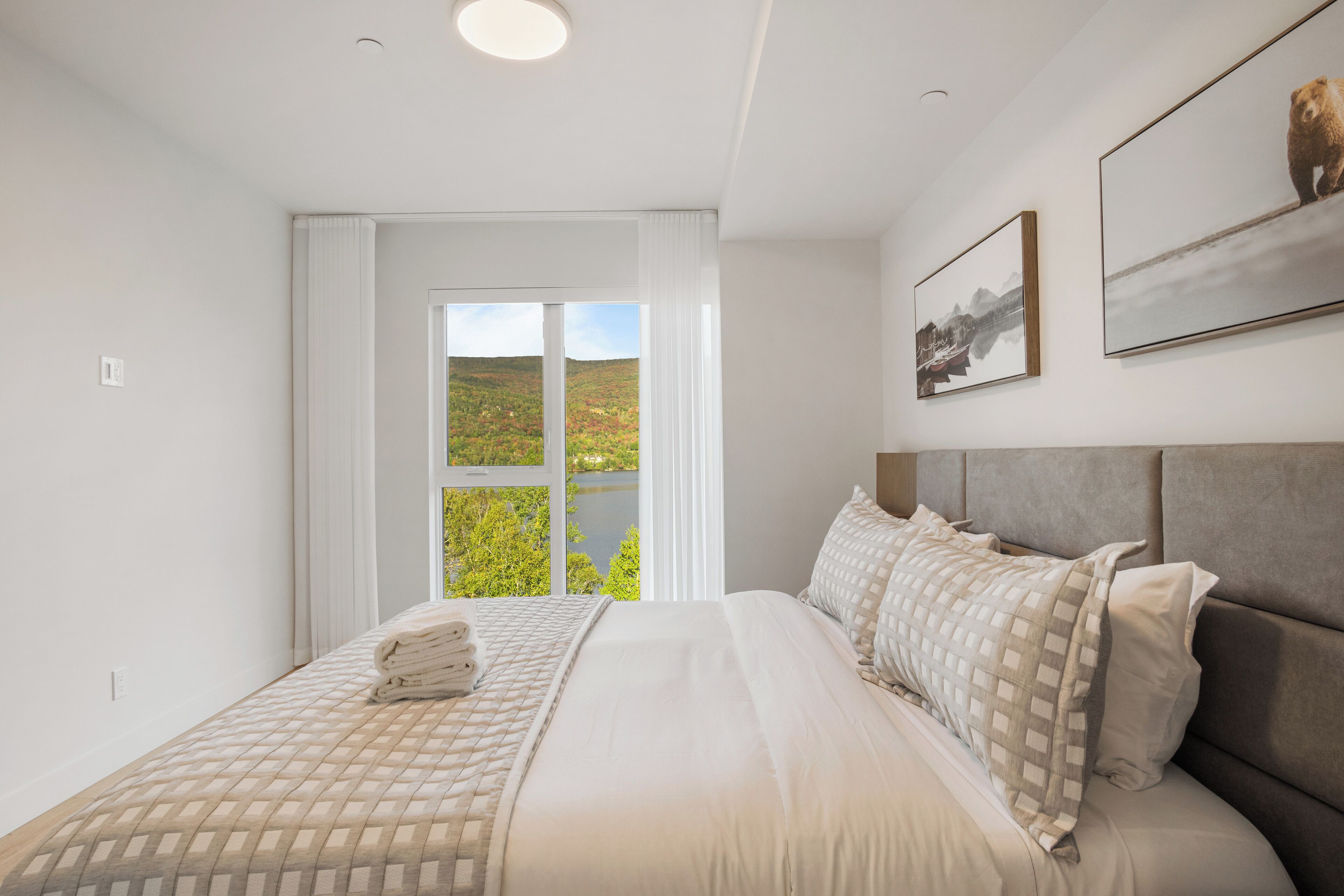 A bedroom with a large bed, a window with a view of greenery, and two framed pictures on the wall.