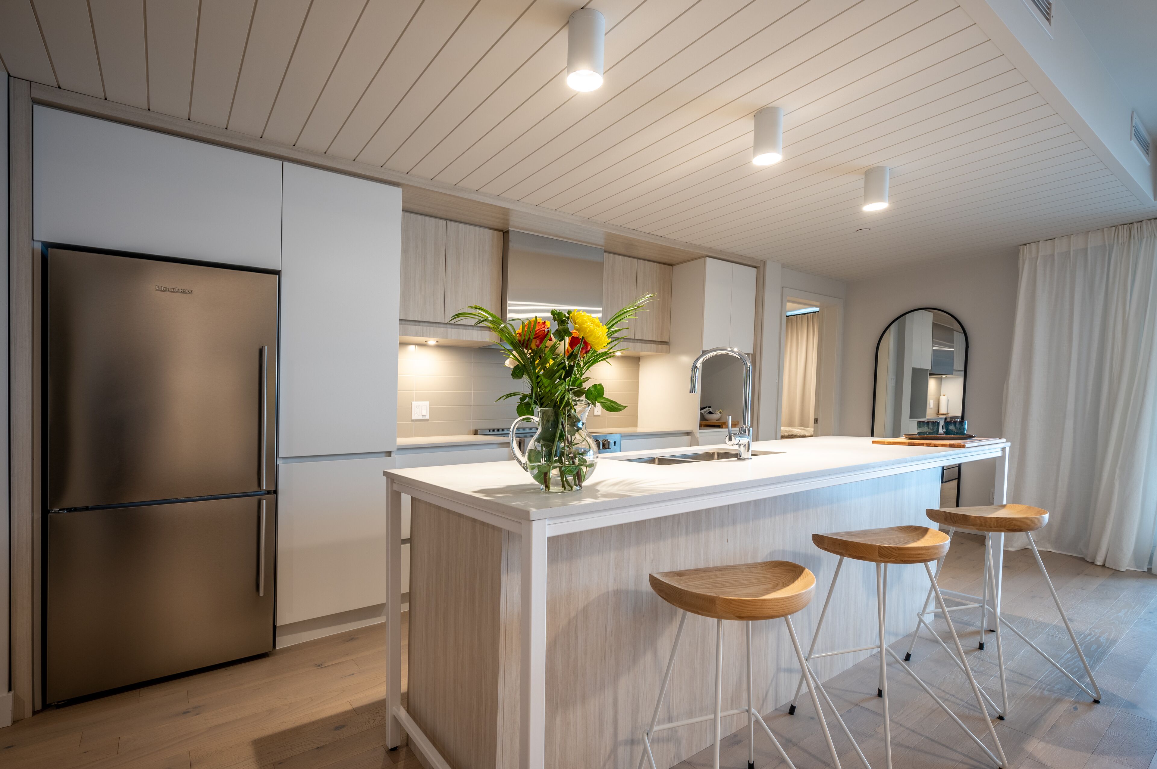 A modern kitchen with a large island, stainless steel refrigerator, and wooden bar stools.