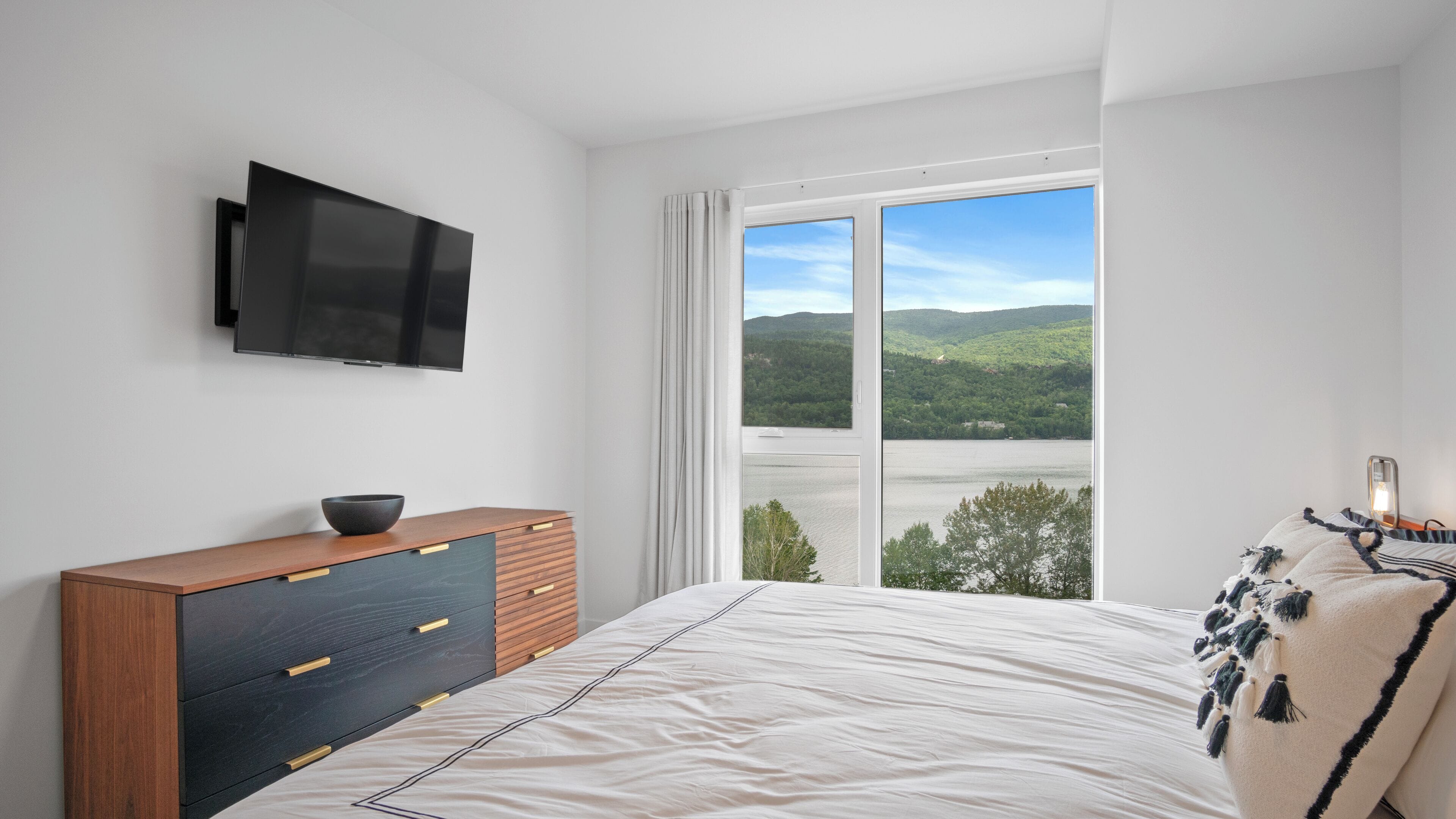 A bedroom with a bed, a TV mounted on the wall, a wooden dresser, and a large window with a view of a lake and hills.