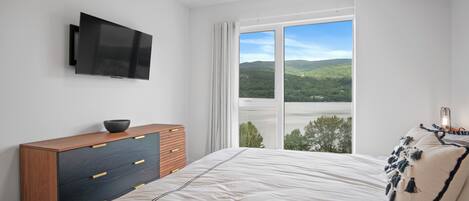 A bedroom with a bed, a TV mounted on the wall, a wooden dresser, and a large window with a view of a lake and hills.