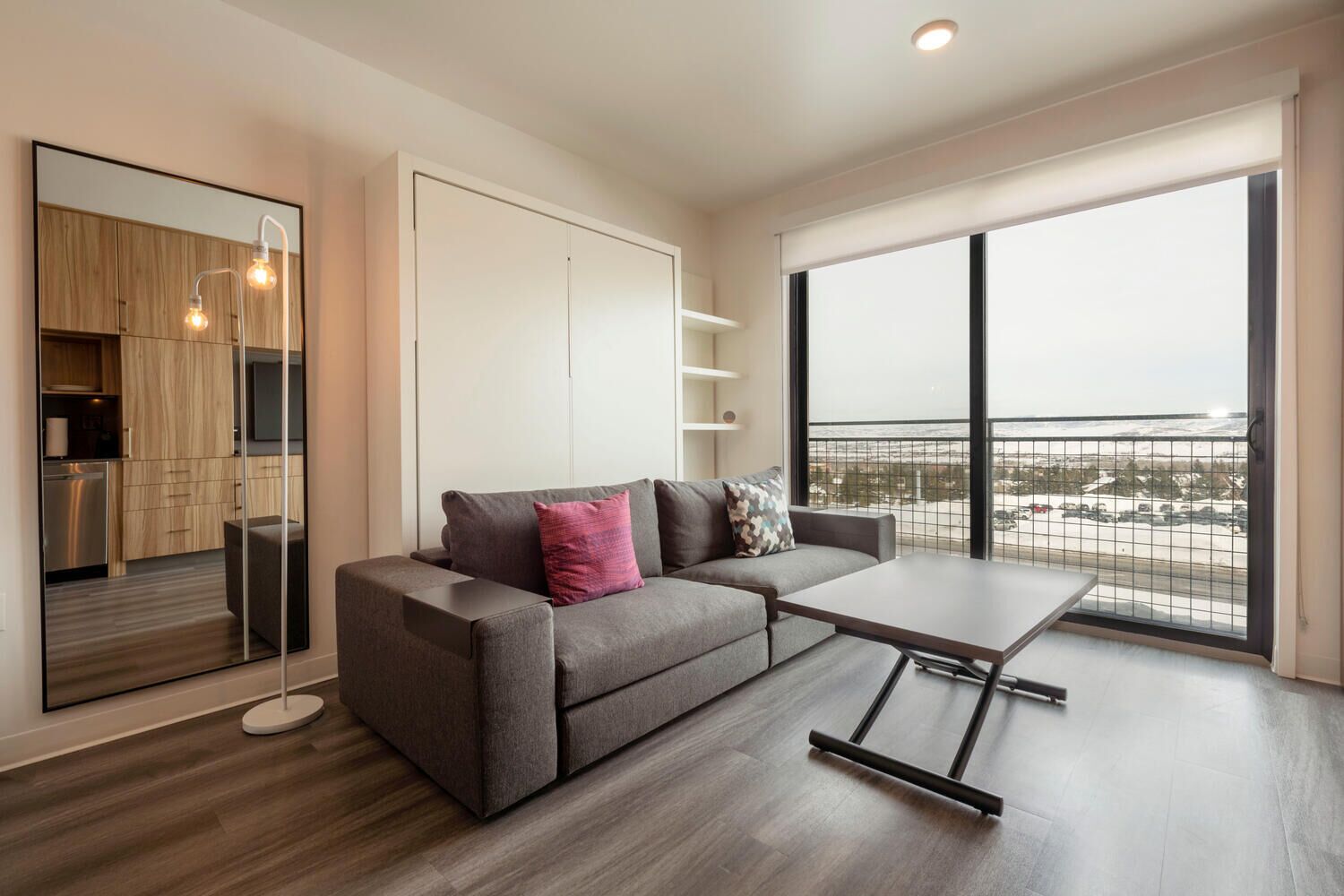 A modern apartment with a grey sofa, a small table, a kitchen with wooden cabinets, and a balcony with a view of a snowy landscape.