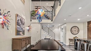 Un vaste espace dédié à la salle à manger, avec une longue table et des chaises modernes, un escalier menant à un niveau supérieur et une grande horloge murale.