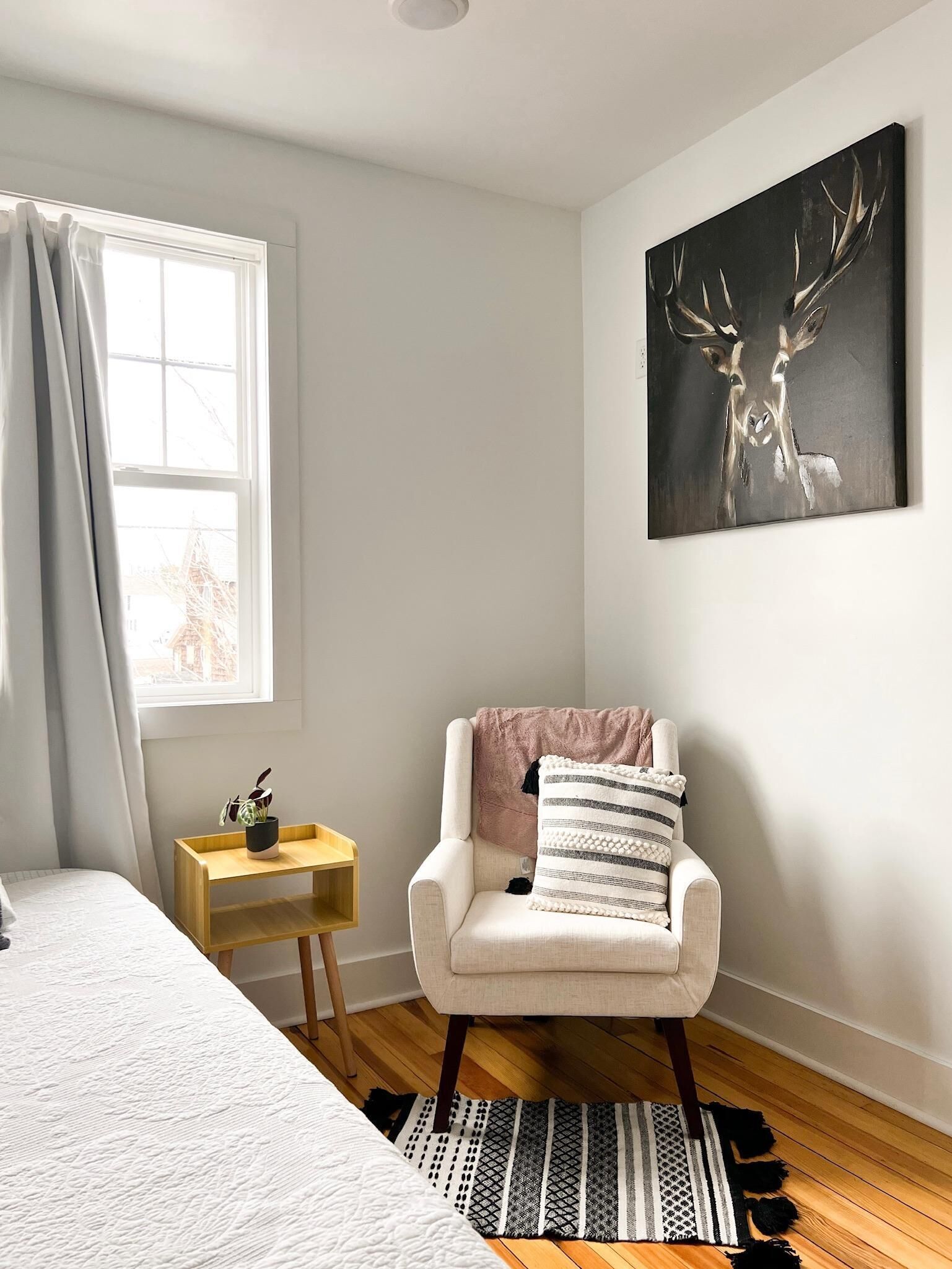 A bedroom with a bed, a chair, a small table, a painting, and a window with curtains.