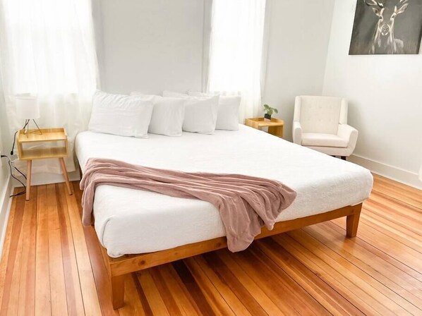 A neatly made bed with a wooden frame and white bedding, a bedside table with a plant, and a chair in a bright room with wooden flooring.