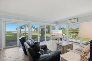 A spacious living area with a leather sofa, a coffee table, and a flat-screen TV. The room has large glass doors leading to a patio with a view of the ocean and palm trees. - Perfect Beachfront Escape at Ocean House 122 (St. Augustine)