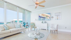 A spacious living room with a large window, a ceiling fan, a glass coffee table, a white sofa, and a decorative chair.