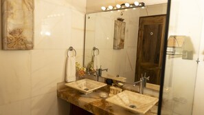 A bathroom with two sinks, a mirror, and a vanity. There are towels hanging on the wall and a shower visible in the reflection.