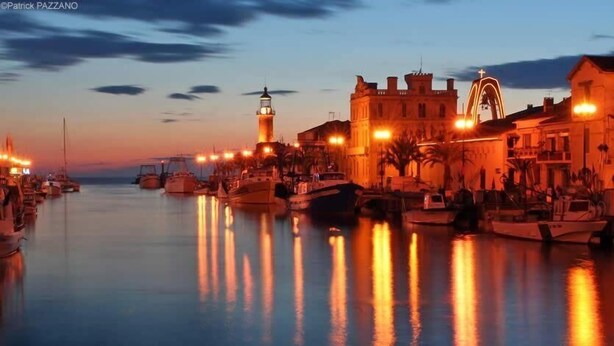 A marina at dusk with boats docked along the waterfront, a prominent building with a clock tower, and a lighthouse in the background.