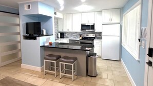 Cocina compacta con armarios blancos, microondas, estufa y refrigerador. Hay una pequeña isla con taburetes y un bote de basura.