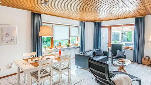 Een moderne woonkamer met een houten plafond, een bank, een eettafel met stoelen en uitzicht op groen buiten.