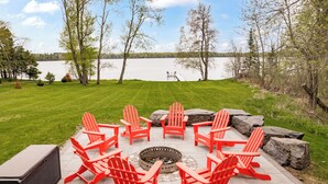 A circular outdoor seating area with red Adirondack chairs around a fire pit, overlooking a lake.