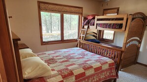 Une chambre avec un grand lit, un lit superposé, une fenêtre donnant sur les arbres et une tête de lit en bois.