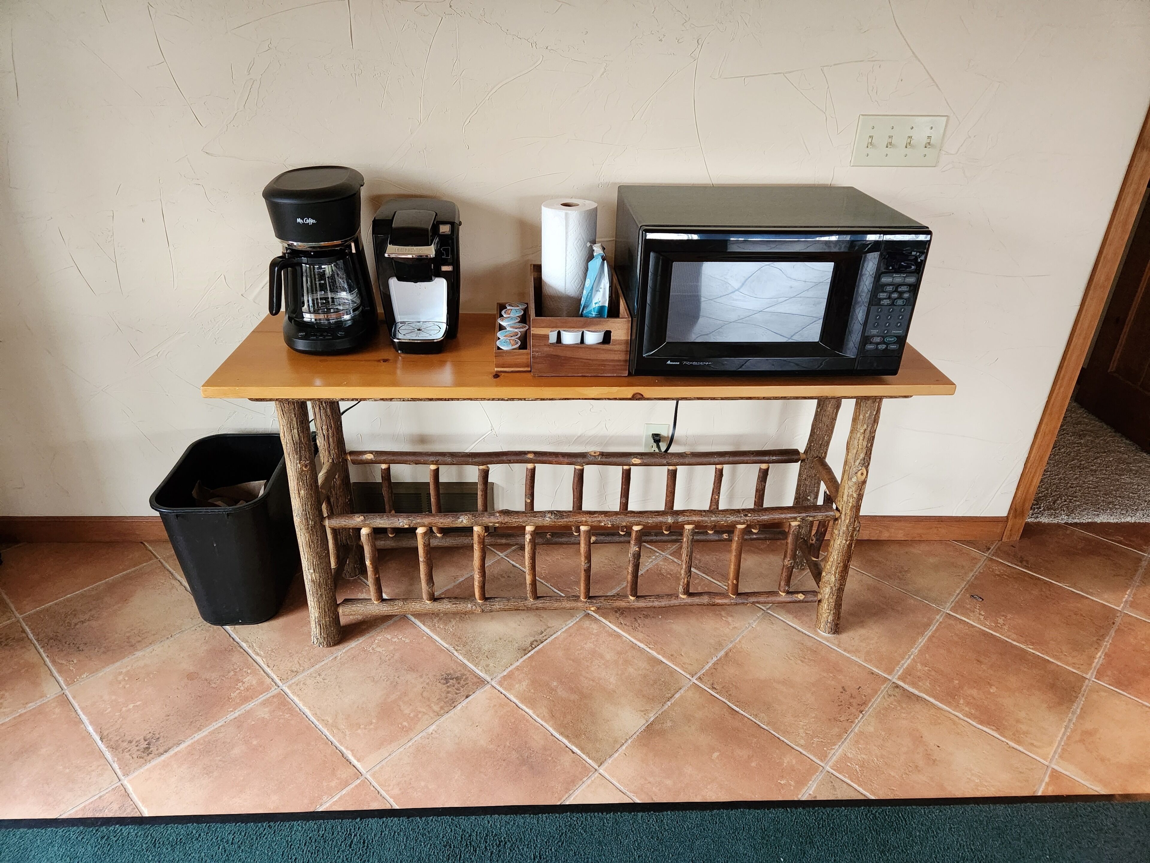 A small kitchen area with a coffee maker, microwave, and a trash bin.