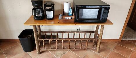A small kitchen area with a coffee maker, microwave, and a trash bin.