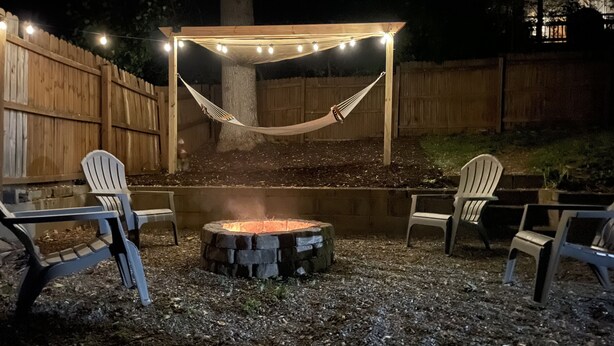 An outdoor patio area with a fire pit, two Adirondack chairs, and a hammock under string lights.