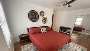 A bedroom with a bed, a small bedside table, a chair, a ceiling fan, and a mirror.