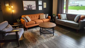 A modern living room with a sofa, armchair, and coffee table, featuring artwork and a view of the outdoors.