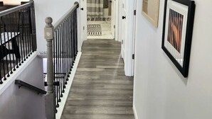 A hallway with a wooden floor, a staircase with black railings, a large wall clock, and a framed picture.