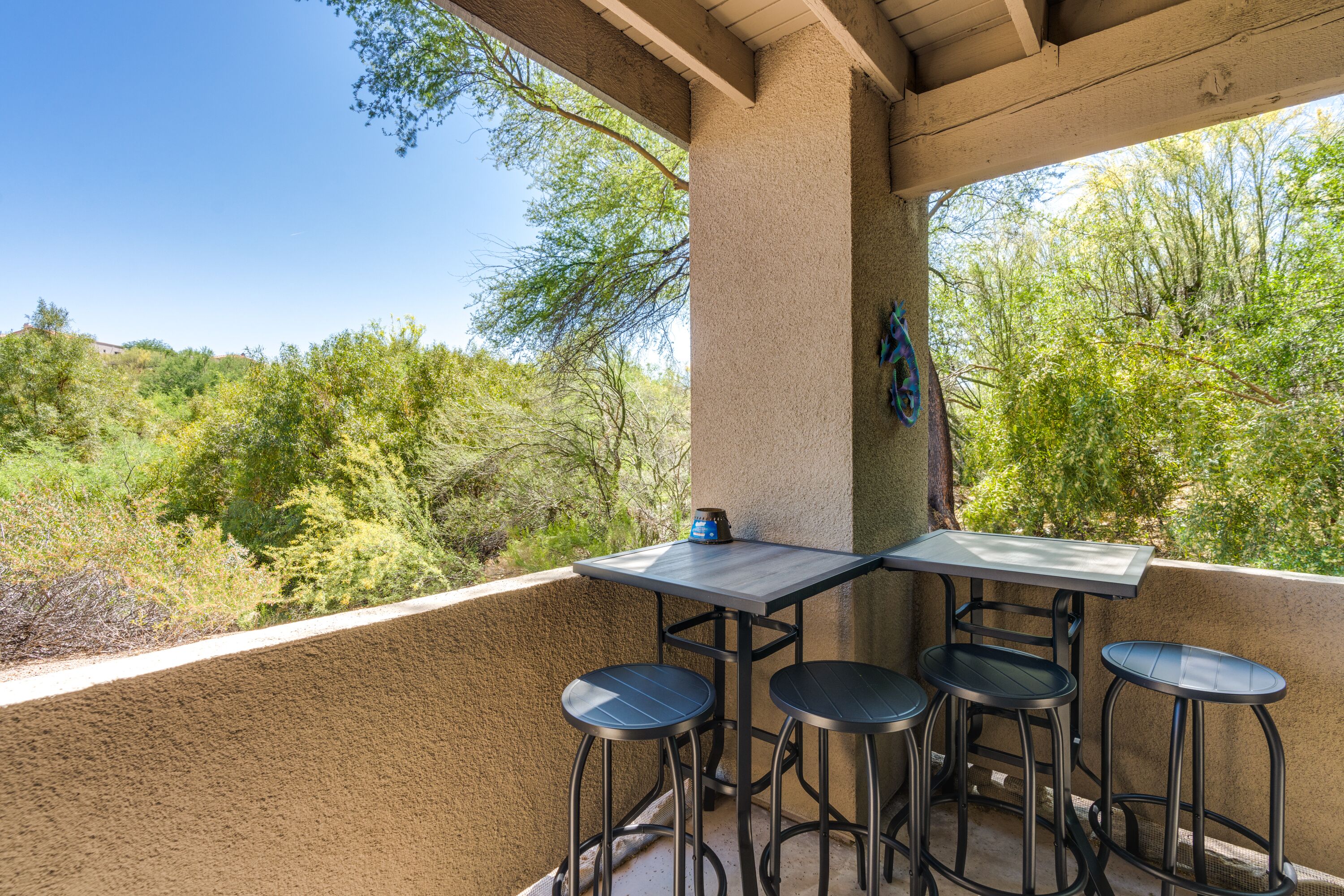 Une terrasse extérieure avec une table et quatre tabourets, entourée d’un mur de couleur beige et donnant sur une zone boisée.