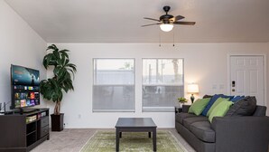 A living room with a dark wooden entertainment center, a flat-screen TV, a coffee table, a gray sofa with green and blue cushions, a potted plant, and a ceiling fan.