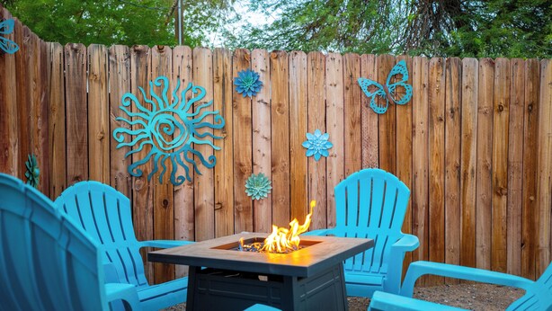 A wooden fence with decorative metal cutouts, a square fire pit with flames, and blue plastic chairs arranged around it.
