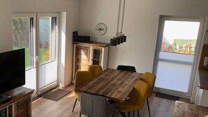 A dining area with a wooden table and yellow chairs, a TV on the left, and a view of greenery outside.