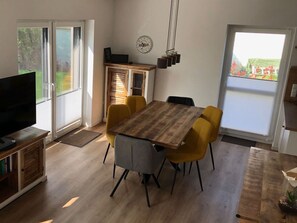 A dining area with a wooden table and yellow chairs, a TV on the left, and a view of greenery outside.