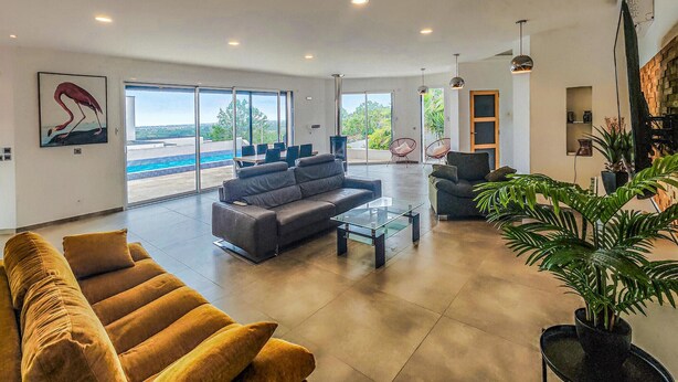 A spacious living room with a gray sofa, a glass coffee table, and a large window offering a view of a swimming pool. - Nice home in Portiragnes (Portiragnes)