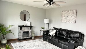 A living room with a fireplace, a black leather sofa, a round mirror, and a large floral artwork on the wall. - Country Getaway, 2- bedroom, Jacuzzi, fire ring (Woodbury)