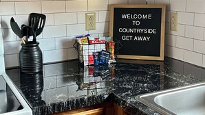A kitchen with wooden cabinets, a marble countertop, a blackboard with a welcome message, and a basket of snacks. - Country Getaway, 2- bedroom, Jacuzzi, fire ring (Woodbury)