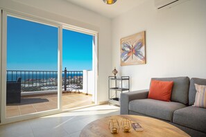 Een woonkamer met een grijze bank, een houten salontafel en uitzicht op zee door een schuifdeur.