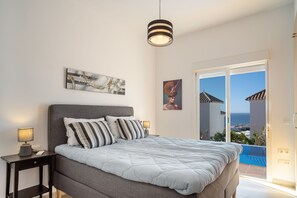 Een slaapkamer met een groot bed, een nachtkastje met een lamp, een kunstwerk aan de muur en uitzicht op zee door een schuifdeur.