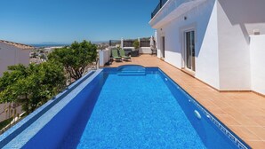 En swimmingpool med udsigt til havet, omgivet af en hvid bygning med balkon.