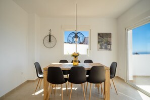 Een eethoek met een houten tafel, zwarte stoelen, een hanglamp en een raam met uitzicht op zee.
