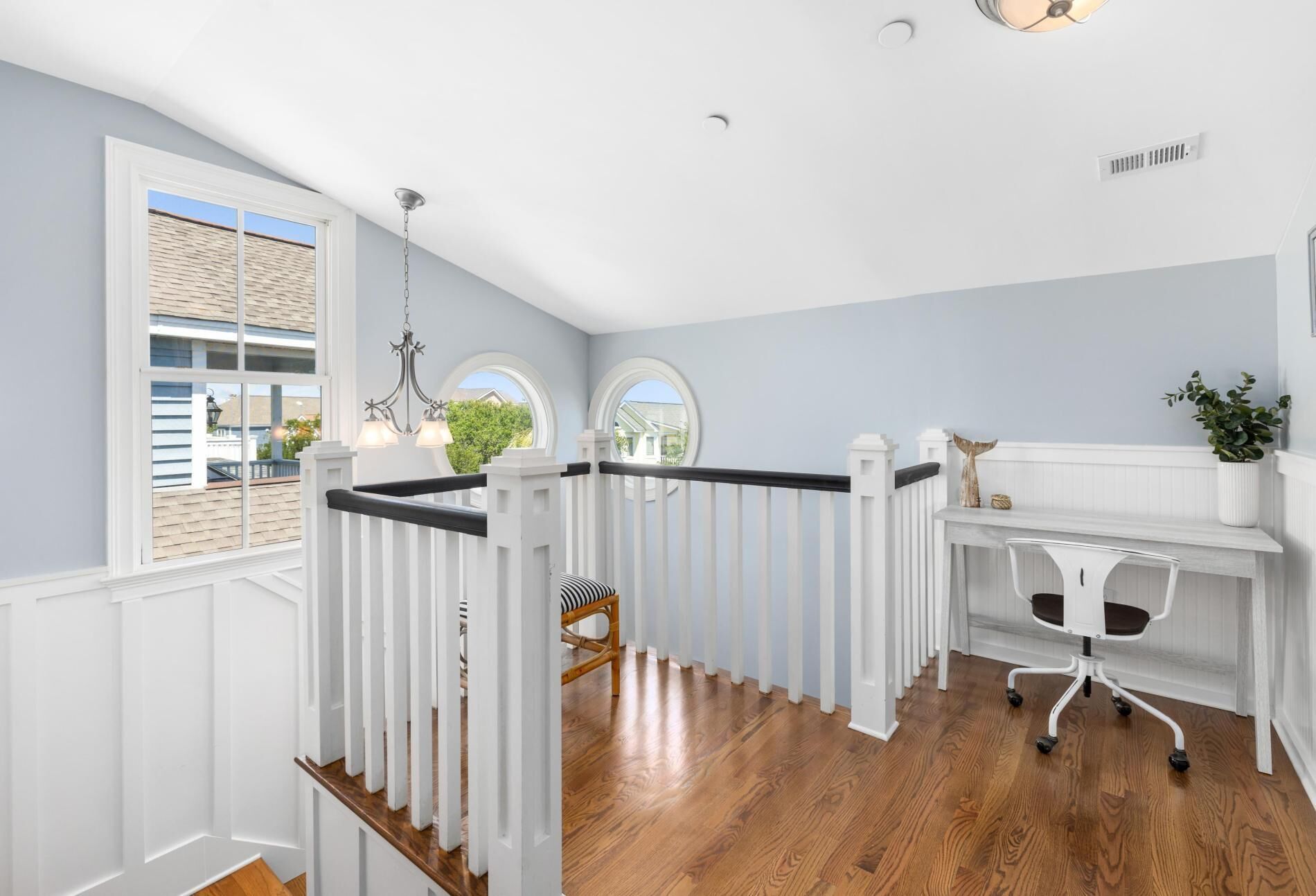 Une pièce avec un plancher en bois, des murs blancs, un bureau blanc avec une chaise, un lustre et une fenêtre donnant sur une maison.