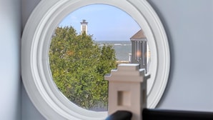 A view through a window with a white frame, overlooking a building with a tower, a body of water, and greenery.
