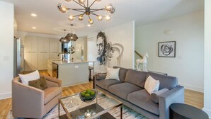 A modern living room with a sofa, armchair, and coffee table. There is a kitchen in the background with cabinets and a dining area. - Midtown Skyline Retreat | Rooftop Deck | Game Room (Nashville)